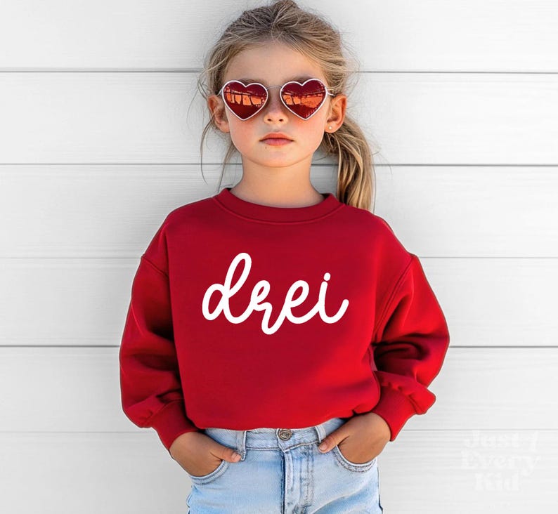 May include: A red sweatshirt with the word "drei" in white script. The child is wearing heart-shaped sunglasses and blue jeans. The sweatshirt has a crew neck and long sleeves.
