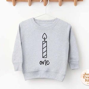 Könnte beinhalten: Hellgraues Sweatshirt mit schwarzer Kerzengrafik und dem Wort "one" darunter. Das Sweatshirt hängt an einem weißen Kleiderbügel vor einem weißen Hintergrund.