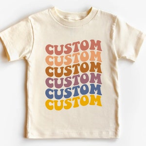 K&ouml;nnte beinhalten: Ein cremefarbenes T-Shirt mit dem Wort "CUSTOM", das sechsmal in einem Regenbogen aus Farben wiederholt wird, darunter Rosa, Orange, Braun, Lila, Blau und Gelb.