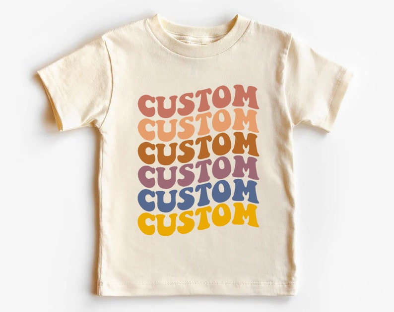 K&ouml;nnte beinhalten: Ein cremefarbenes T-Shirt mit dem Wort "CUSTOM", das sechsmal in einem Regenbogen aus Farben wiederholt wird, darunter Rosa, Orange, Braun, Lila, Blau und Gelb.