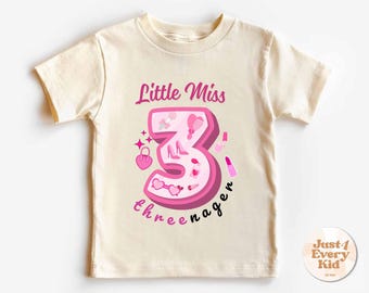 May include: Cream-colored t-shirt with a pink graphic design for a three-year-old. The design includes the words "Little Miss 3neenager" and features pink illustrations of a purse, high heels, lipstick, and sunglasses.