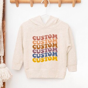 K&ouml;nnte beinhalten: Ein hellbeiger Kapuzenpullover mit dem Wort "CUSTOM", das sechsmal in einem Regenbogen aus Farben wiederholt wird, darunter Rot, Orange, Gelb, Gr&uuml;n, Blau und Lila.