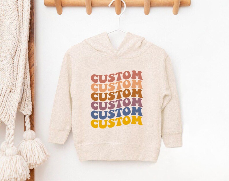 K&ouml;nnte beinhalten: Ein hellbeiger Kapuzenpullover mit dem Wort "CUSTOM", das sechsmal in einem Regenbogen aus Farben wiederholt wird, darunter Rot, Orange, Gelb, Gr&uuml;n, Blau und Lila.
