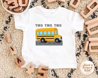 Peut inclure: T-shirt blanc avec un dessin de bus scolaire jaune et le texte "TWO TWO TWO". Le bus a des pneus noirs et une bande noire. Le t-shirt est entouré de blocs de bois et d'un tapis.
