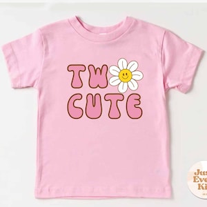 May include: A pink toddler t-shirt with the words "TWO CUTE" in brown outlined letters and a smiling daisy graphic. The shirt is made of a soft material and is designed for children.