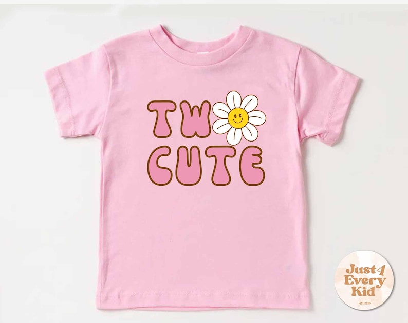 May include: A pink toddler t-shirt with the words "TWO CUTE" in brown outlined letters and a smiling daisy graphic. The shirt is made of a soft material and is designed for children.