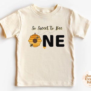Può includere: T-shirt color crema per bambini piccoli con grafica a tema api. Il design include un alveare giallo e marrone, due api dei cartoni animati e la scritta "So Sweet To Bee ONE" in nero. La parola "ONE" è in lettere grandi e in grassetto.