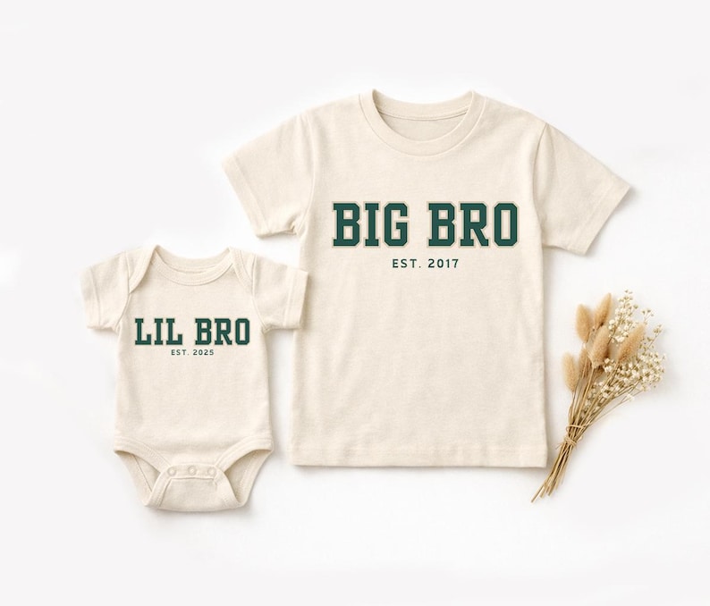 Puede incluir: Un body de manga corta color crema con el texto verde oscuro LIL BRO EST. 2025 y una camiseta de manga corta a juego color crema con el texto verde oscuro BIG BRO EST. 2017, ambos con un dise&ntilde;o de fuente de estilo universitario.
