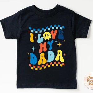 Może przedstawiać: Czarny T-shirt z kolorowym retro wzorem, na kt&oacute;rym widnieje napis "Kocham mojego tatę" z uśmiechniętą buźką.