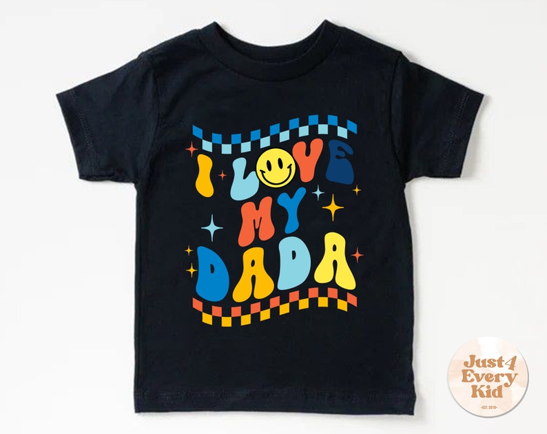 Może przedstawiać: Czarny T-shirt z kolorowym retro wzorem, na kt&oacute;rym widnieje napis "Kocham mojego tatę" z uśmiechniętą buźką.