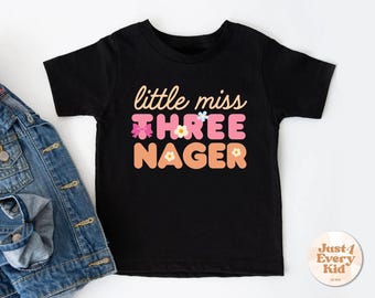 May include: Black toddler t-shirt with the text "little miss THREE NAGER" in a retro font. The word "THREE" is in pink, and "NAGER" is in orange. The shirt has floral accents and is next to a denim jacket.