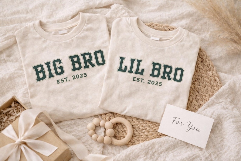 Puede incluir: Dos camisetas de cuello redondo color blanco roto con letras de estilo universitario en verde oscuro. Una camiseta muestra el texto BIG BRO EST. 2025 y la otra LIL BRO EST. 2025. Las camisetas est&aacute;n dispuestas con un mordedor de madera y una caja de regalo.