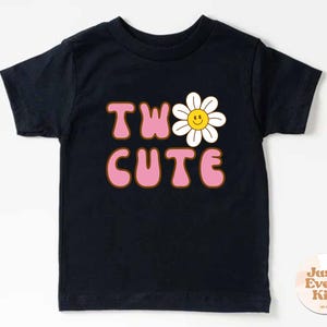 May include: A black t-shirt with the words "TWO CUTE" in pink bubble letters with a brown outline. A white daisy with a yellow smiling face replaces the "O". The shirt is for kids.