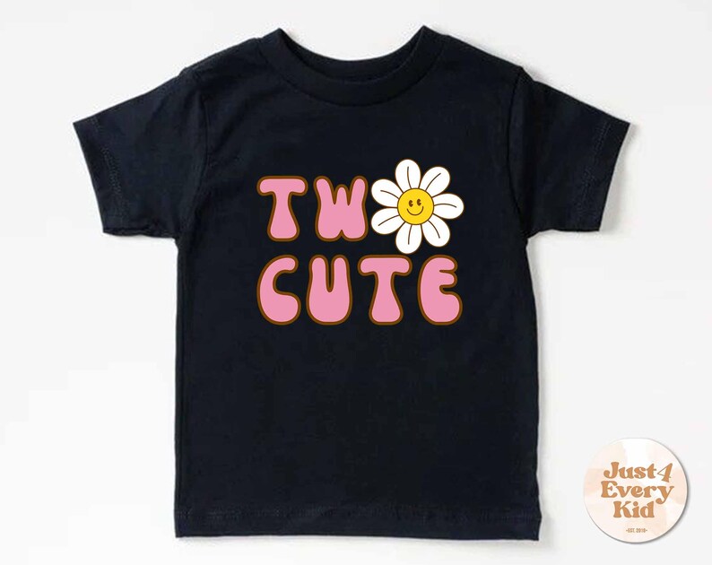 May include: A black t-shirt with the words "TWO CUTE" in pink bubble letters with a brown outline. A white daisy with a yellow smiling face replaces the "O". The shirt is for kids.