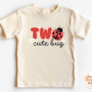 Puede incluir: Camiseta crema para niños pequeños con las palabras "TWO cute bug" en rojo y negro. Un gráfico de mariquita está al lado de la palabra "TWO". Pequeñas flores blancas están colocadas alrededor del texto. La camiseta es de manga corta y cuello redondo.
