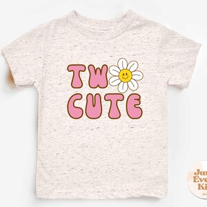 May include: A light gray t-shirt with the words "TWO CUTE" in pink bubble letters with a brown outline. A smiling daisy graphic is next to the word "TWO". The shirt has a round neck and short sleeves.