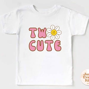 May include: White t-shirt with the words "TW0 CUTE" in pink bubble letters with a brown outline. A smiling daisy graphic is next to the word "TW0". The shirt has short sleeves and a round neck. A small circular logo is in the bottom right corner.