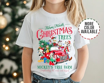 Puede incluir: Camiseta blanca con un diseño navideño. Presenta a Mickey Mouse y sus amigos frente a un camión rojo, con el texto "Farm Fresh Christmas Trees" y "Mickey's Tree Farm". La camiseta también dice "Complementary hot cocoa and beignets".