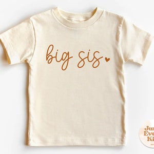 Puede incluir: Una camiseta de manga corta de color crema con el texto "big sis" en letra cursiva marrón con un pequeño corazón.