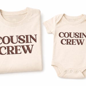 May include: Two cream-coloured shirts with the text "COUSIN CREW" in brown. One is a folded t-shirt, and the other is a baby onesie. The items are displayed on a white background.