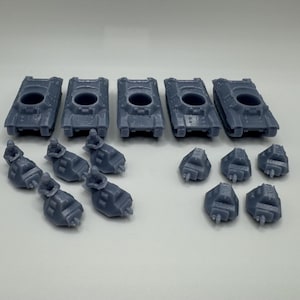 May include: A collection of small, gray toy tanks and other military vehicles. The tanks have a rectangular shape with a circular opening on top. There are also smaller, detailed pieces that appear to be turrets or other components. The items are arranged on a white surface.