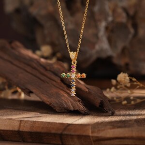 Op de afbeelding: Een gouden ketting met een kleurrijk kruis als hanger. Het kruis is versierd met kleine, glinsterende edelstenen in verschillende kleuren, waaronder rood, blauw, groen en geel.