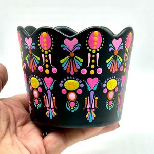 May include: A black ceramic planter with a scalloped rim, adorned with a vibrant, repeating pattern of pink, yellow, orange, and teal floral designs. The planter is held in a hand against a white background.