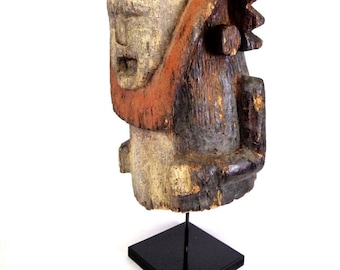 May include: A wooden sculpture of a human head with a stylized face and a red and black painted design. The sculpture is mounted on a black stand.