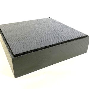 Può includere: Una scatola quadrata di legno nero con coperchio. La scatola ha una finitura a venature del legno strutturata. Il coperchio è a filo con i bordi della scatola. La scatola è su sfondo bianco.