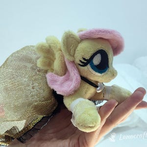 May include: A plush toy pony with yellow fur, pink accents, and blue eyes. The pony is wearing a gold mesh dress with black trim. The pony is a children's toy.