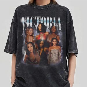 May include: Dark gray t-shirt featuring a group of women with the word "VICTORIA" in white block letters. The design includes a lightning bolt graphic. The shirt has a vintage, distressed look.