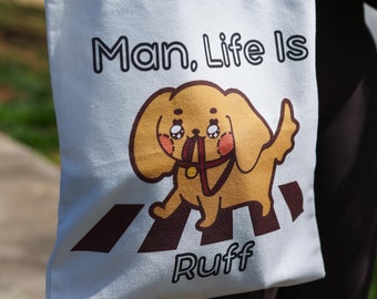 Könnte beinhalten: Weiße Canvas-Tragetasche mit einem Cartoon-Hund, der eine rote Leine trägt, und dem aufgedruckten Text "Man. Life Is Ruff".