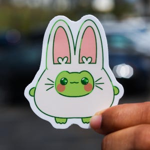May include: A green frog wearing a white bunny costume with pink ears. The frog has large, round eyes and a small, pink nose.