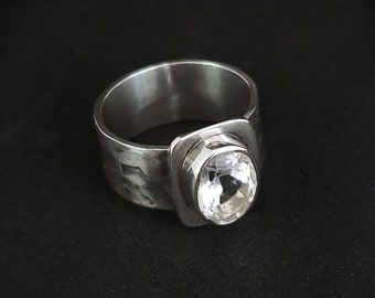 May include: A silver ring with a large, clear, oval-shaped gemstone set in a square bezel. The band of the ring is textured and has a hammered finish.