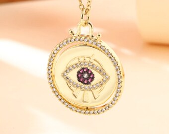 May include: Gold necklace with a round pendant featuring a detailed evil eye design with pink and white stones. The pendant is surrounded by a circle of smaller white stones.