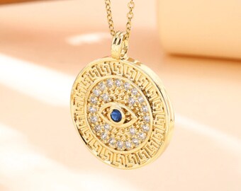 May include: Gold necklace with a round pendant featuring a blue evil eye surrounded by clear rhinestones and a Greek key design.