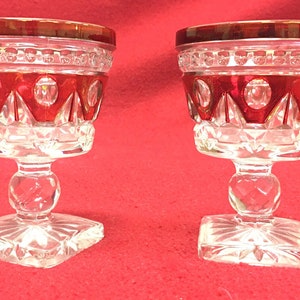 May include: Two clear glass goblets with a red rim and a cut glass design. The goblets have a square base and a round stem.
