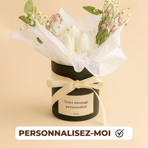 Peut inclure: Un coffret rond noir, orné d'un ruban crème, contenant des fleurs blanches et de la verdure. Le coffret porte une étiquette avec le texte "Votre message personnalisé" et le texte "PERSONNALISEZ-MOI" en dessous. Fond beige uni.
