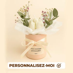 Könnte beinhalten: Ein hellrosa zylindrischer Behälter, gebunden mit einem cremefarbenen Band, gefüllt mit weißen tulpenförmigen Kerzen und frischen Blumen. Der Behälter hat ein Etikett mit der Aufschrift "Votre message personnalise". Der untere Teil des Bildes zeigt "PERSONNALISEZ-MOI".