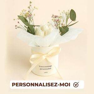 May include: A cream-coloured cylindrical gift box tied with a ribbon, holding white tulip-shaped candles and greenery. The box features the text "Votre message personnalisé" and "PERSONNALISEZ-MOI".