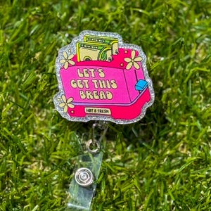 May include: A pink and silver glitter badge reel with the text "LET'S GET THIS BREAD" and a graphic of a cash register with money inside. The badge reel has a clear plastic clip and a retractable cord.