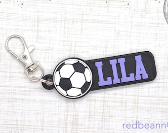 以下が含まれることがあります： 黒と白のサッカーボールのキーホルダーで、紫色のネームタグには「LILA」と書いてあります。