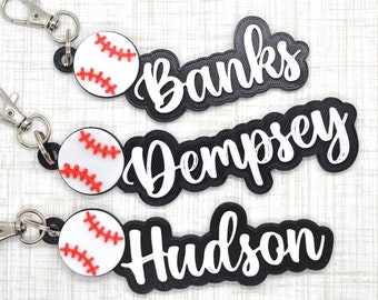 May include: Three baseball-themed keyrings with silver clasps. Each keyring features a white baseball with red stitching and a black nameplate with a white name: Banks, Dempsey, and Hudson. The keyrings are on a light wood-grain surface.