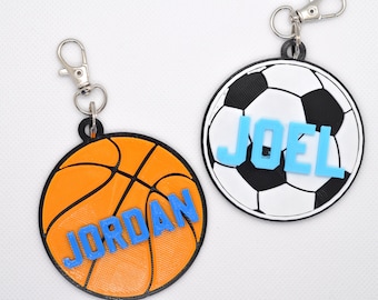 May include: Two personalized sports-themed keychains. One keychain features a black and white soccer ball with the name "JOEL" in blue letters. The other keychain features an orange basketball with the name "JORDAN" in blue letters.