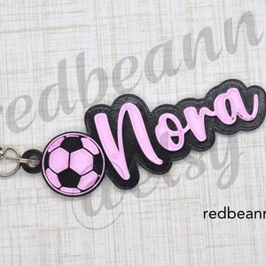 May include: Black and pink personalized keychain with the name "Nora" and a pink soccer ball charm.