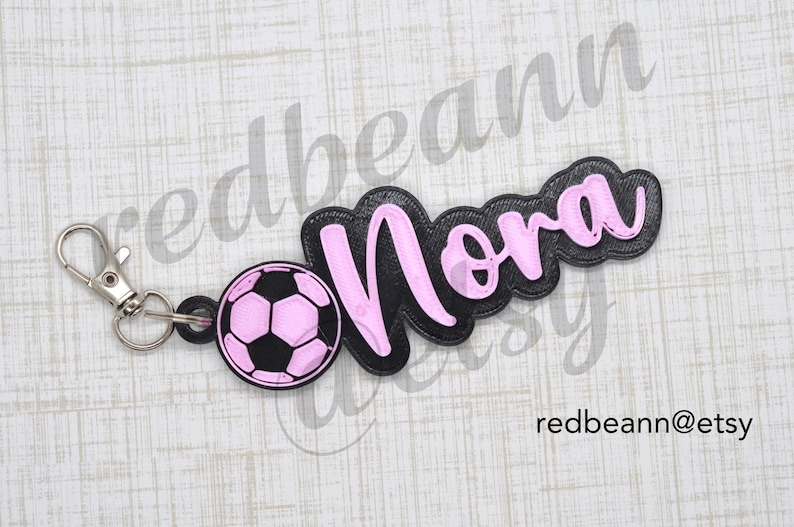 May include: Black and pink personalized keychain with the name "Nora" and a pink soccer ball charm.