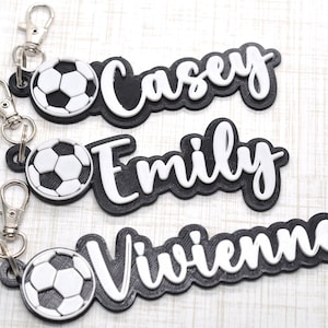 May include: Three black and white personalized keychains with a soccer ball charm. The names Casey, Emily, and Vivienne are written in white on the black keychains.