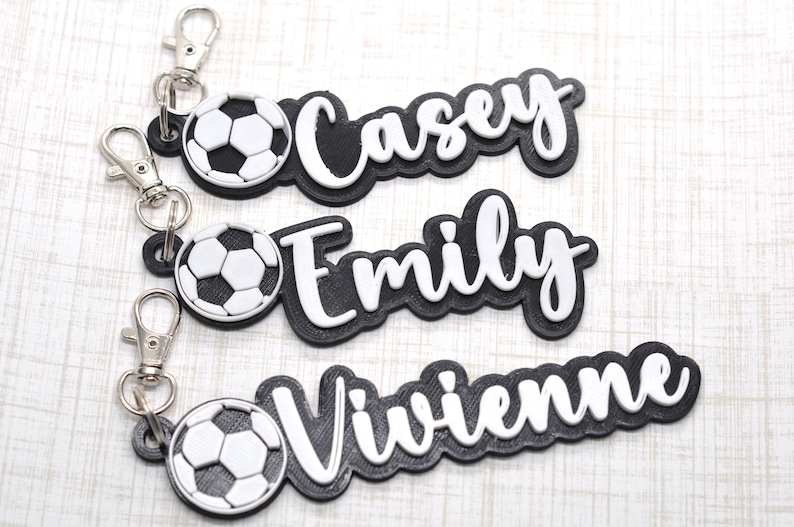 May include: Three black and white personalized keychains with a soccer ball charm. The names Casey, Emily, and Vivienne are written in white on the black keychains.