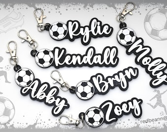 以下が含まれることがあります： 「Rylie」、「Kendall」、「Molly」、「Brynn」、「Abby」、「Zoey」という名前とサッカーボールが描かれた、黒と白のパーソナライズされたサッカーボールキーホルダー7個。