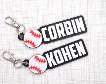 Puede incluir: Dos llaveros de béisbol personalizados en blanco y negro con llaveros plateados. Las etiquetas tienen los nombres "CORBIN" y "KOHEN" impresos en letras blancas. Cada etiqueta tiene una pelota de béisbol blanca con costuras rojas.